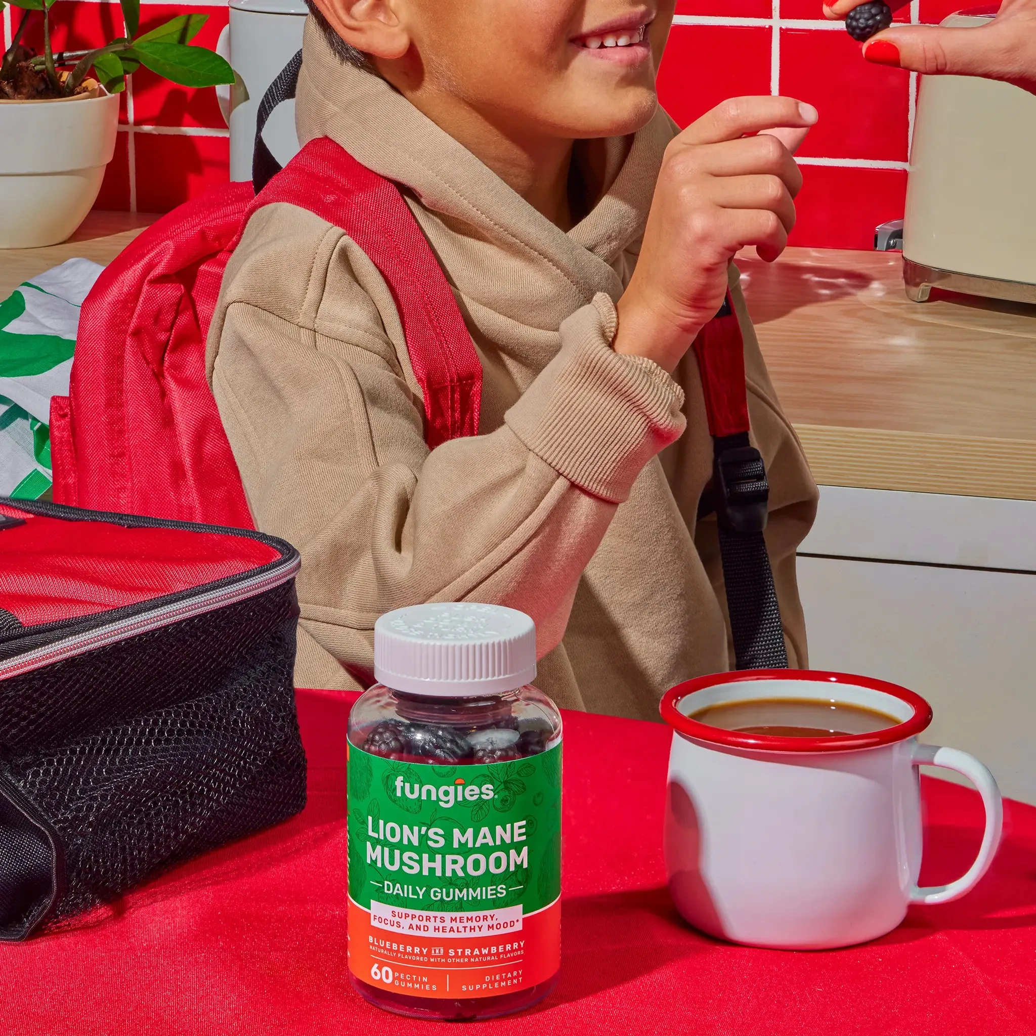 Child with red backpack at table with Lion's Mane Mushroom Gummies bottle, beside a mug and lunch bag, accepting a berry. Gummies support cognitive performance.